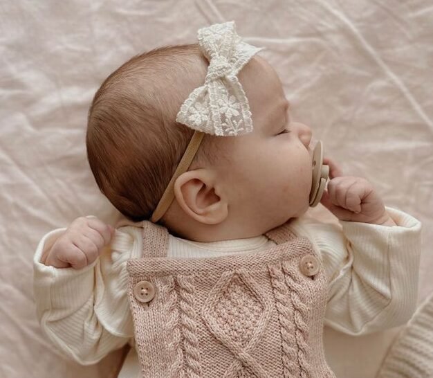 The baby sleeps soundly, cheeks rosy and peaceful, with a tiny bow resting atop their soft hair. Each breath is slow and sweet, wrapped in a world of warmth and innocence.