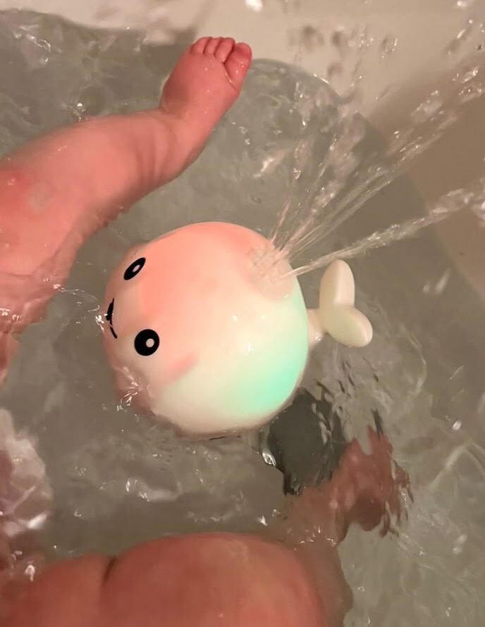 Baby giggles as he stretches his tiny hands toward the floating whale toy, his fingers just brushing the glowing surface before it sprays water, making him squeal with excitement and splash even more.