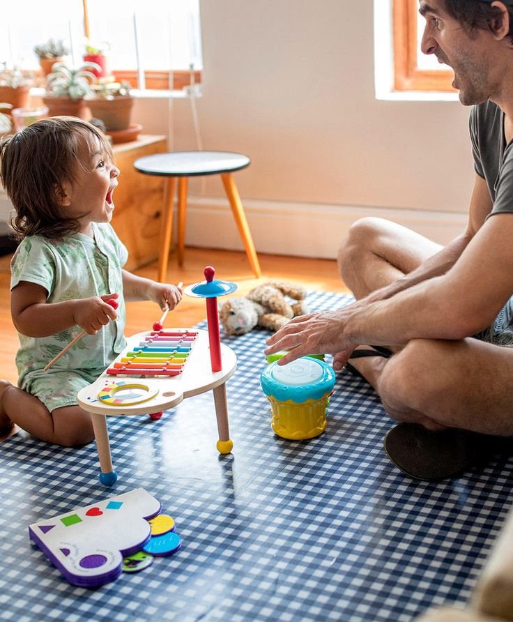 the baby girl joyfully taps on a small musical instrument, her tiny hands guided by her father’s loving touch. They share a moment of laughter and connection, as the father encourages her to explore the sounds. It’s a beautiful, playful bond between parent and child, filled with music, love, and learning.